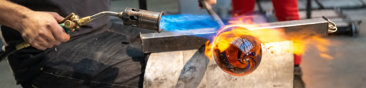 Man holds blow torch to glass art piece that is fixed to the end of a glass blowing stick.