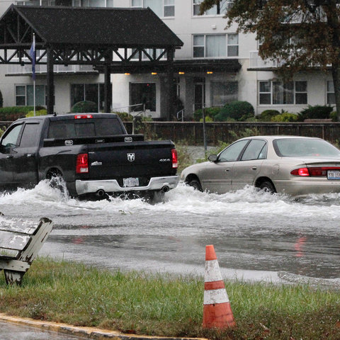 a black pick-up truck and a white sedan drive through a flooded street