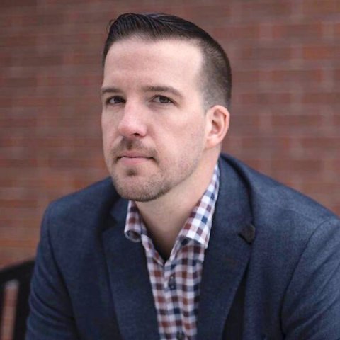 Larson dressed in a blue suit standing in front of brick wall. 
