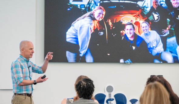 Douglas Casa stands in a lecture hall gesturing with his hands towards a screen