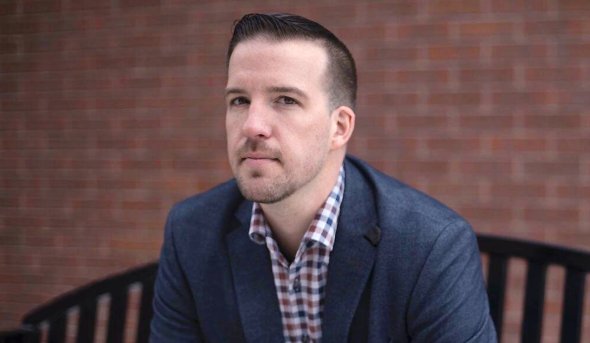 Larson dressed in a blue suit standing in front of brick wall. 