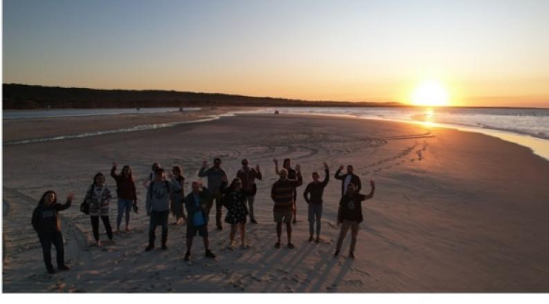 2022 Ocean Hack Week participants wave on the beach as the sun sets