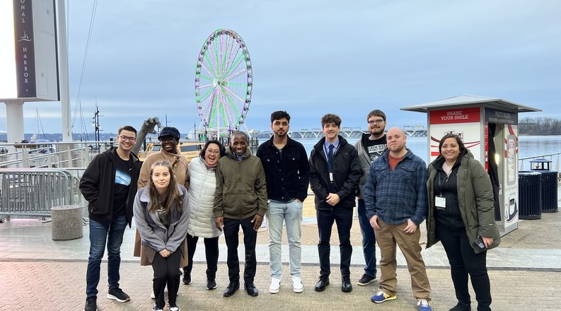 A group of students stand on a sidewalk in front of the National Harbor in Maryland. 