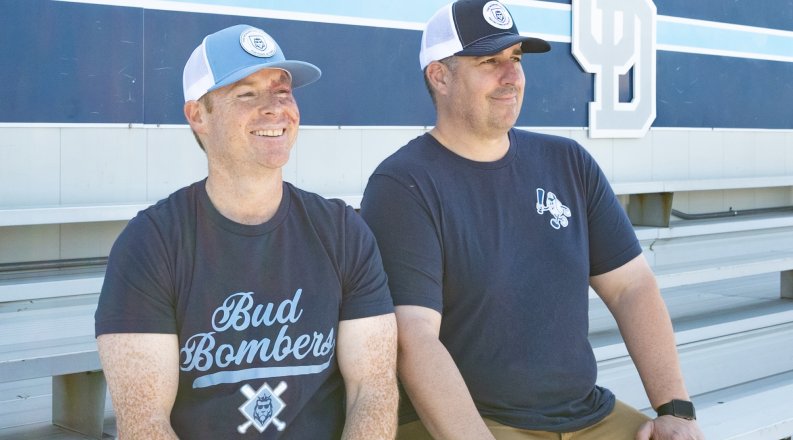 two men in blue shirts and white and blue caps sit on bleachers