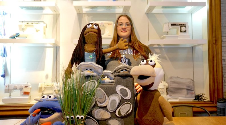 Sydney Walker poses with puppet characters from “The Amazing Neighborhood Mollusk Adventure” including Mr. Blue the crab, Reign the mermaid, a trio of oysters called the Reef Rascals and Esperanza, a brown pelican. Two clumps of marsh grass with googly eyes, named Tina and Flora, stand in the foreground.