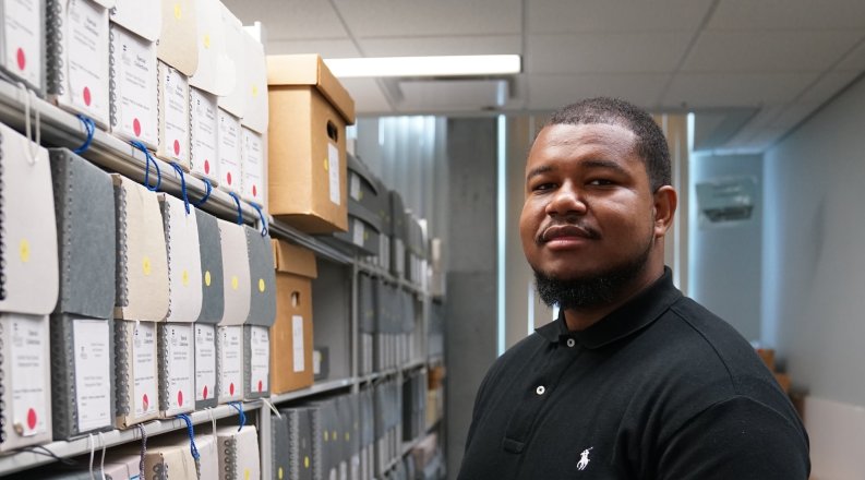 A student stands next to a bookshelf in a library.