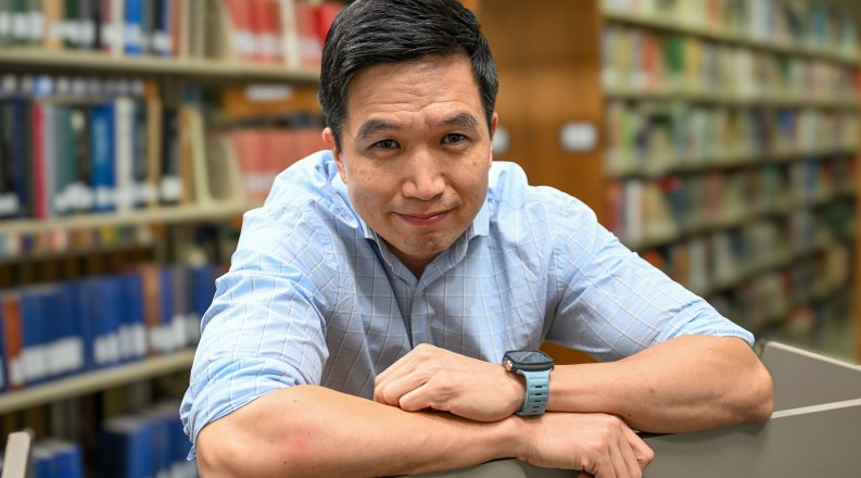 John Park, Ph.D., looks into the camera, leaning with arms crossed on bookshelf in a library.