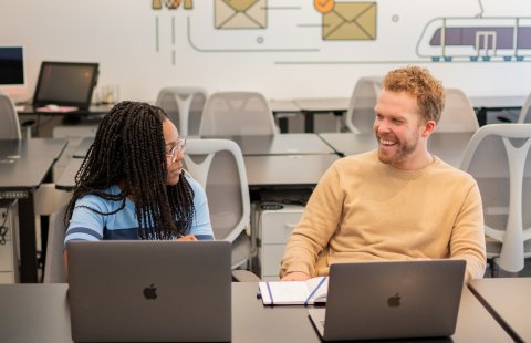 A female student laughs with a male supervisor in an office space while they look at laptops.