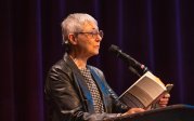 A woman reads from a book on a stage.