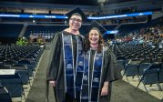 Two people in graduation attire stand in an area decorated for commencement.