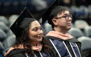 Two people in graduation attire, including caps and robes, smile during a graduation ceremony.