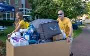 Two people push a cart full of items into a dorm.