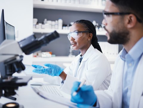 Man and woman in laboratory wearing lab coats with microscope and computers.