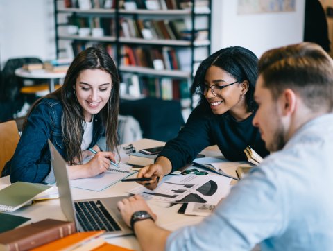 Three students watching educational video on laptop in library, skilled team of young people using netbook and wifi in library