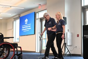 A man walking assisted by a woman and a cane