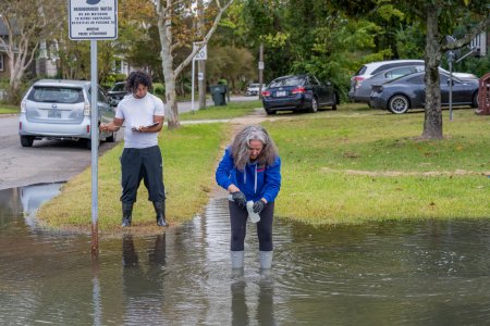A female scientist bends down to collect a water sample from a flooded street.
