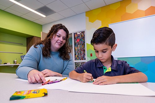 A female therapist in blue shirt sits with a boy as he color