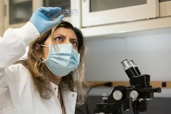 A masked woman is looking up at a microscope slide. Blue mat